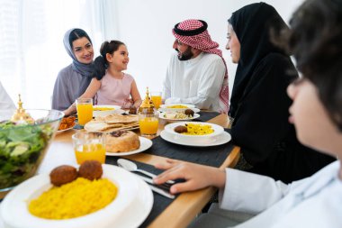 Suudi Arabistan kıyafeti giyen mutlu Arap ailesi akşam yemeği sırasında evde birlikte vakit geçiriyorlar - Orta Doğu Suudi aileleri ve çocukları Ramazan 'ı gün be gün kutluyorlar.