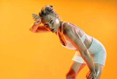 Güzel, olgun bir kadın spor kıyafeti giyiyor spor yapıyor. Terli, olgun bir sporcu kadın yoğun bir egzersizden sonra duruyor. Alnını canlı turuncu bir arka planda kararlılıkla siliyor.