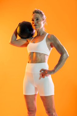 Spor kıyafetleri giyen güzel, olgun bir kadın spor yapıyor. Spor salonunda turuncu arka planda egzersiz yaparken beyaz spor giysisi giymiş, tıp topu kaldıran kendine güvenen bir kadın.