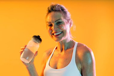 Spor kıyafetleri giyen güzel, olgun bir kadın spor yapıyor. Mutlu, sportif, gri saçlı, beyaz spor sutyen takan, elinde su şişesi tutan, spor ve sağlık durumunu kutlayan olgun bir kadın.