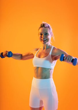 Spor kıyafetleri giyen, spor giyinen güzel olgun bir kadın. Formda, olgun bir kadın halterlerini turuncu renkli bir arka plana doğru kaldırıyor. Egzersiz yaparken kendine güveni, gücü ve canlılık saçıyor.