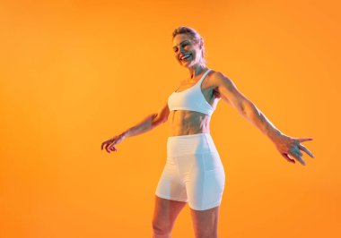 Spor kıyafetleri giyen güzel, olgun bir kadın spor yapıyor. Kollarını açmış sevinçle poz veriyor. Mutlu. Beyaz spor giyimli formalı son sınıf öğrencisi canlı turuncu bir arkaplan karşısında duruyor.