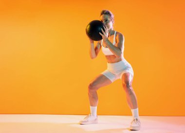 Spor kıyafetleri giyen güzel olgun bir kadın spor yapıyor. Güçlü bir bayan sporcu. Stüdyo ortamında canlı turuncu bir arka planda sağlık topu ile çömelme hareketleri yapıyor.