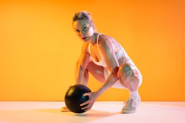 Spor kıyafetleri giyen güzel olgun bir kadın spor yapıyor. Kaslı olgun bir kadın canlı turuncu arka plana karşı ilaç topuyla çömelme hareketleri yapıyor. Formda ve güçlü olduğunu gösteriyor.