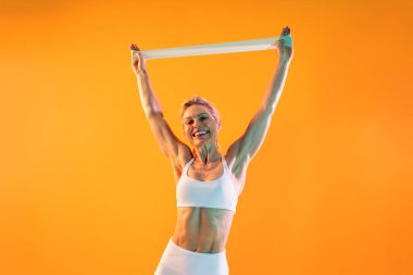 Güzel, olgun, spor kıyafetleri giyen spor giyinen bir kadın spor yapıyor. Mutlu bir son sınıf spor modeli. Bir direniş bandını kaldırıyor, gülümsüyor ve canlı turuncu bir spor salonunun arka planında kaslarını gösteriyor.