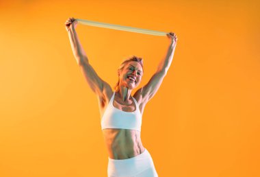 Spor kıyafetleri giyen, spor giyinen, güzel, olgun bir kadın spor salonunda turuncu bir arka planda elastik bir bantla spor yaparken mutlu bir spor modeli eğitimi alıyor, kaslı kaslarını gösteriyor ve gülümsüyor.