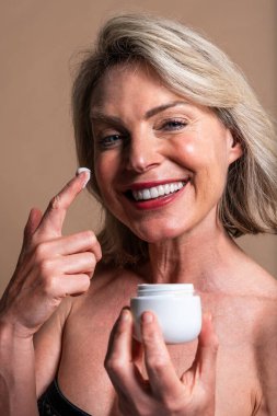 Güzel olgun bir kadının güzel portresi, kozmetik, cilt bakımı ve sağlıkla ilgili kavramlar. Kısa sarı saçlı, gülümseyen olgun bir kadın beyaz bir kavanozdan yüz kremi sürüyor.