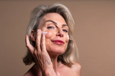 Güzel olgun bir kadının güzel portresi, kozmetik, cilt bakımı ve sağlıkla ilgili kavramlar. Gülümseyen olgun bir kadın yüzüne nemlendirici sürüyor. Sağlıklı olmak için cilt bakımını benimsemek