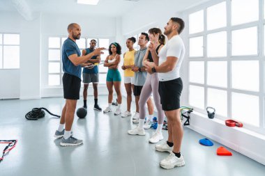 Spor salonlarında spor yapan çok ırklı bir grup sporcu, spor salonlarında spor yapan bir grup sporcunun antrenörlerini dinleyerek spor salonunda spor aletleriyle antreman yapmalarını sağlıyor.