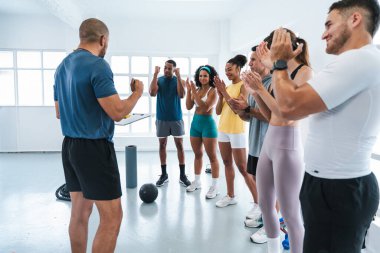 Çok ırklı spor yapan bir grup sporcu, modern bir jimnastik dalında işlevsel bir antrenmanı tamamladıktan sonra antrenman yapan çeşitli sporcuları alkışlıyor ve koçlarına tezahürat yapıyorlar.