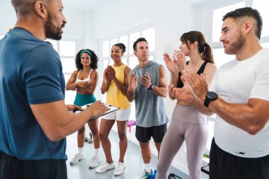 Spor salonunda egzersiz yapan çok ırklı bir grup sporcu, spor salonundaki çalışmalarını bitirdikten sonra antrenörlerini alkışlıyorlar.