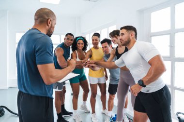 Spor salonlarında spor yapan çok ırklı bir grup sporcu, antrenörleri bir spor salonunda notlar alırken, spor salonundaki sporcular da elele tutuşuyor.