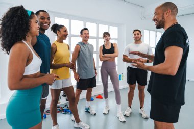 Beden eğitimi sınıfında spor yapan çok ırklı bir grup sporcu, spor salonundaki egzersizleri açıklayan spor eğitmenlerini dinleyen gülümseyen sporcular.