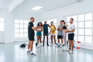 Çok ırklı spor yapan bir grup sporcu spor salonunda antrenörlerini dinleyen çok ırklı gülümseyen sporculardan oluşan bir grup parlak bir spor salonunda yapılan işlevsel bir egzersizi anlatıyor.