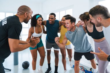 Spor salonunda egzersiz yapan çok ırklı bir grup sporcu spor salonundaki antrenörleriyle spor salonundaki spor başarılarını kutluyorlar.