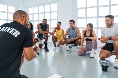 Spor salonunda spor yapan çok ırklı bir grup sporcu antrenörlerini dinleyen gülümseyen çok ırklı sporcular parlak bir spor salonunda yeni işlevsel eğitim tatbikatlarını anlatıyor.