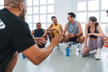 Spor salonunda egzersiz yapan çok ırklı bir grup sporcu jimnastik yapıyor. Jimnastik salonunun zemininde oturan çeşitli atletlere jest yapıyor ve onlarla konuşuyor.