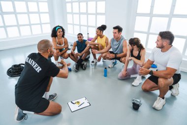 Spor salonunda spor yapan çok ırklı bir grup sporcu spor salonunda çok ırklı bir grup sporcu spor salonunda oturuyorlar ve antrenörlerinin yaptıkları bir egzersiz hakkında konuştuklarını dinliyorlar.