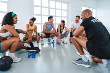 Spor salonunda spor yapan çok ırklı bir grup sporcu spor salonunda gülümseyen bir grup sporcu spor salonunun zemininde daireler çizerek oturuyorlar.