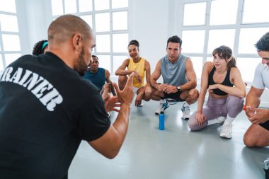 Spor salonunda spor yapan çok ırklı bir grup sporcu spor salonunda spor salonunda egzersiz yapan sporcu, spor salonunda oturup spor salonunun talimatlarını özümseyen bir grup sporcuya işlevsel egzersizleri anlatıyor.