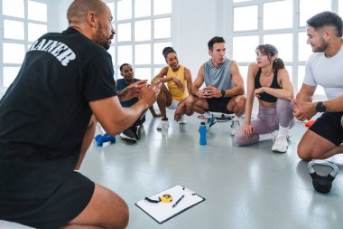 Spor salonunda spor yapan çok ırklı bir grup sporcu spor salonunda spor salonunda spor salonunun zemininde oturan sporculara egzersiz yaptıklarını anlatıyor.