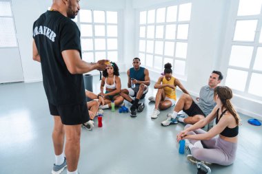 Spor salonunun zemininde oturan çok ırklı sporculardan oluşan bir grup beden eğitimi sınıfında egzersiz yapıyor.