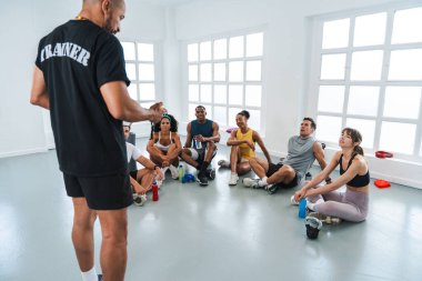 Spor salonunda egzersiz yapan çok ırklı bir grup sporcu spor salonunda antrenörlerini dinleyip dinleniyorlar.