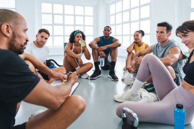 Çok ırklı spor yapan bir grup sporcu beden eğitimi sınıfında çeşitli sporcular antrenörleriyle aynı çemberde oturuyorlar.