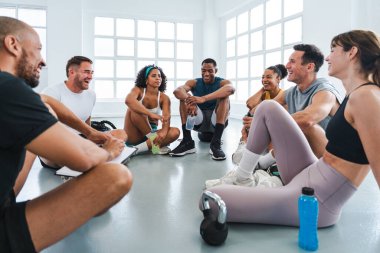 Spor salonunda spor yapan çok ırklı bir grup sporcu spor salonunda gülümseyen sporcular spor salonunun zemininde bir çember etrafında oturuyorlar.