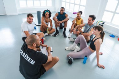 Spor salonunda spor yapan çok ırklı bir grup sporcu spor salonunda gülümseyen sporcular spor salonunun zemininde bir çember etrafında oturuyorlar.