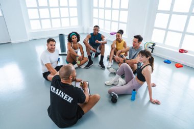 Spor salonunun zemininde bir çember içinde oturan çok ırklı sporculardan oluşan bir grup beden eğitimi sınıfında egzersiz yapıyor.