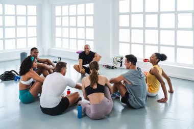 Spor salonunda spor yapan çok ırklı bir grup sporcu spor salonunun zemininde çember şeklinde dinlenen çok ırklı sporculara gülümsüyor.