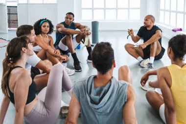 Spor salonunun zemininde bir çember içinde oturan çeşitli sporcuların eğitim sınıfında egzersiz yapan çok ırklı bir grup. Form hedefleri için takım çalışması ve iletişim teşvik ediliyor