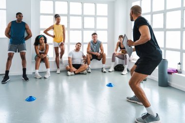 Çok ırklı spor yapan bir grup sporcu, beden eğitimi sınıfında çeşitli sporculardan oluşan bir grupta, kişisel antrenörlerin fonksiyonel eğitim sırasında düzgün bir şekilde çömelme tekniğini göstermelerini izliyorlar.