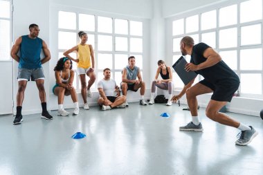Spor salonunda spor yapan çok ırklı bir grup sporcu, özel antrenörlerini dinleyip parlak bir spor salonunda egzersiz yapıyor.