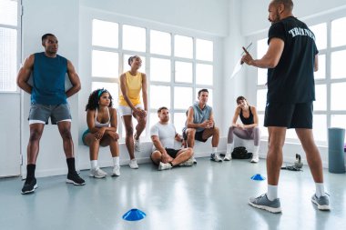 Çok ırklı spor yapan bir grup sporcu, antrenörlerini dinleyen çok ırklı sporculardan oluşan bir spor salonunda egzersiz yapıyorlar.