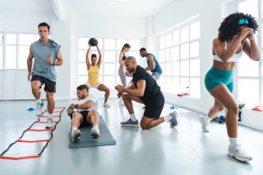 Çok ırklı spor yapan bir grup sporcu, parlak bir spor salonunda antrenman yapan farklı sporcularla antrenman yapıyor.