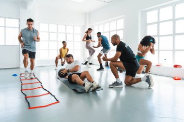 Çok ırklı spor yapan bir grup sporcu, bir tıp topuyla egzersiz yapan çok ırklı sporculardan oluşan bir beden eğitimi grubunda çalışıyor. Çeviklik merdiveni