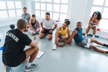 Çok ırklı spor yapan bir grup sporcu, parlak bir spor salonunda antrenman yaparken antrenörlerini dinleyen çok ırklı sporculardan oluşan bir beden eğitimi grubunda çalışıyor.