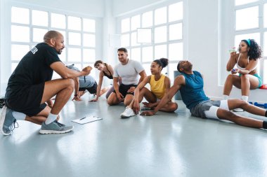Çok ırklı spor yapan bir grup sporcu, farklı sporculardan oluşan bir beden eğitimi grubunda antrenörlerini dinleyip parlak bir spor salonunda egzersiz yapıyorlar.