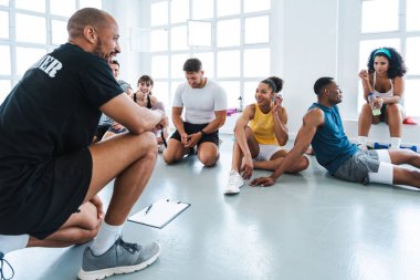 Çok ırklı bir grup sportif insan, antrenman sınıfında spor yapıyor. Eğitmenlik yapıyor ve elinde bir pano tutuyor.