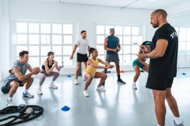 Çok ırklı spor yapan bir grup sporcu beden eğitimi sınıfında çok ırklı sporcular, fonksiyonel bir eğitim sırasında kişisel antrenörün rehberliğinde çömelme hareketleri yapıyorlar.
