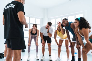 Çok ırklı bir grup sporcu spor salonunda spor yapıyor. Spor salonunda spor yaparken gülümseyen sporcular antrenörlerini dinliyor. Parlak bir spor salonunda.