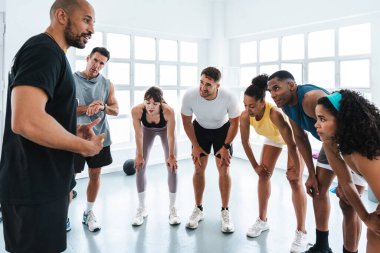 Spor salonunda egzersiz yapan çok ırklı bir grup sporcu antrenörlerinin parlak bir spor salonunda yapacağı bir sonraki işlevsel egzersizi anlatırken yorgun sporcular eğiliyor.