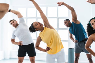 Antrenman sınıfında çeşitli sporcuları çalıştıran çok ırklı bir grup sporcu yan esneme hareketleri yapıyor.