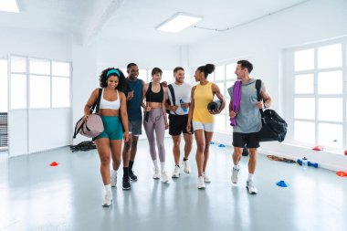 Çok ırklı spor yapan bir grup sporcu spor salonunda egzersiz yapıyor. Fonksiyonel bir antrenmandan sonra gülümseyen sporcular yürüyorlar ve sohbet ediyorlar. Spor çantaları taşıyorum. Yoga paspasları