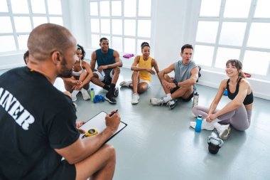 Çok ırklı spor yapan bir grup sporcu, antrenman sınıfında spor yapıyor. Bir not panosuna not alıyor. Fonksiyonel eğitim sırasında gülümseyen atletlerle konuşuyor.