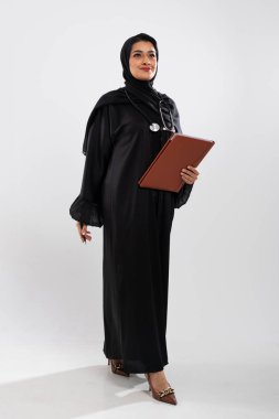 Geleneksel abaya ve tesettür giyen, elinde bir dosya ve steteskop tutan, stüdyo ortamında sağlık ve profesyonelliği temsil eden kendinden emin Müslüman kadın doktor.