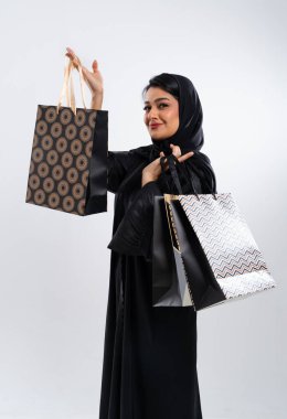 Geleneksel siyah abaya ve tesettür takan Arap kadın, elinde beyaz bir stüdyo geçmişine karşı alışveriş poşetleri tutarken gülümsüyor ve modern tüketim ve kültür mirasının bir karışımını sergiliyor.
