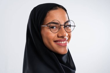 Geleneksel siyah başörtüsü ve zarif yuvarlak gözlükler takan neşeli bir ortadoğulu kadının stüdyo portresi, temiz beyaz bir arka plana karşı özgüvenini ve modern tarzını sergiliyor.